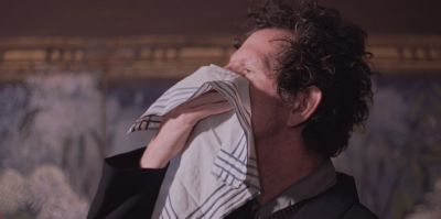 Film still from 'Great Noses Are Blown Alike' (2024) by Jon Beaton. A man is poised to blow his nose into a white handkerchief with decorative blue lines around the edges.